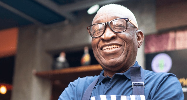 Man with a big smile is wearing thick, black glasses, a blue shirt, and a striped apron.